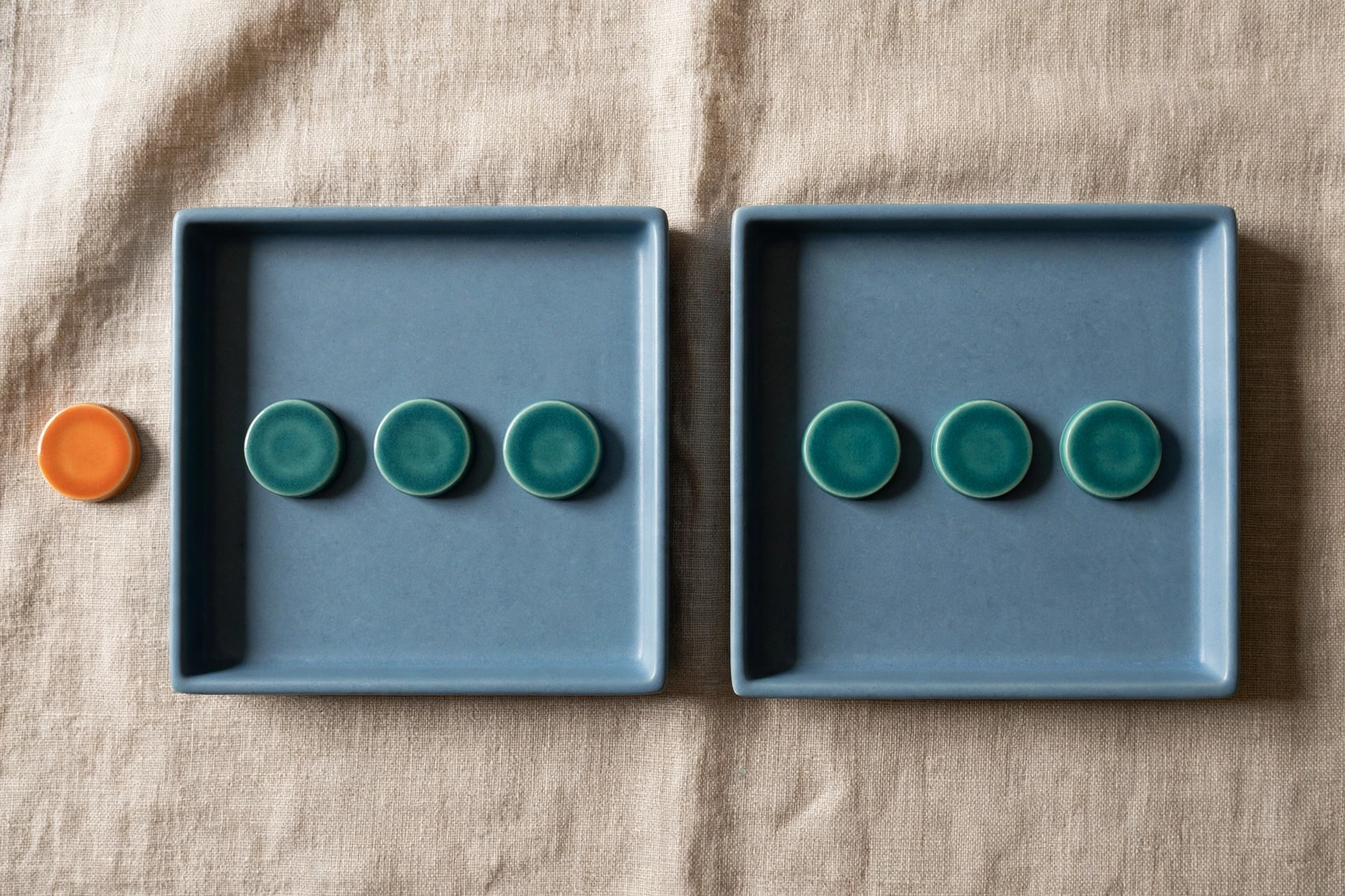 Two blue trays each hold three teal tokens, with one orange token placed beside the left tray on a neutral fabric.