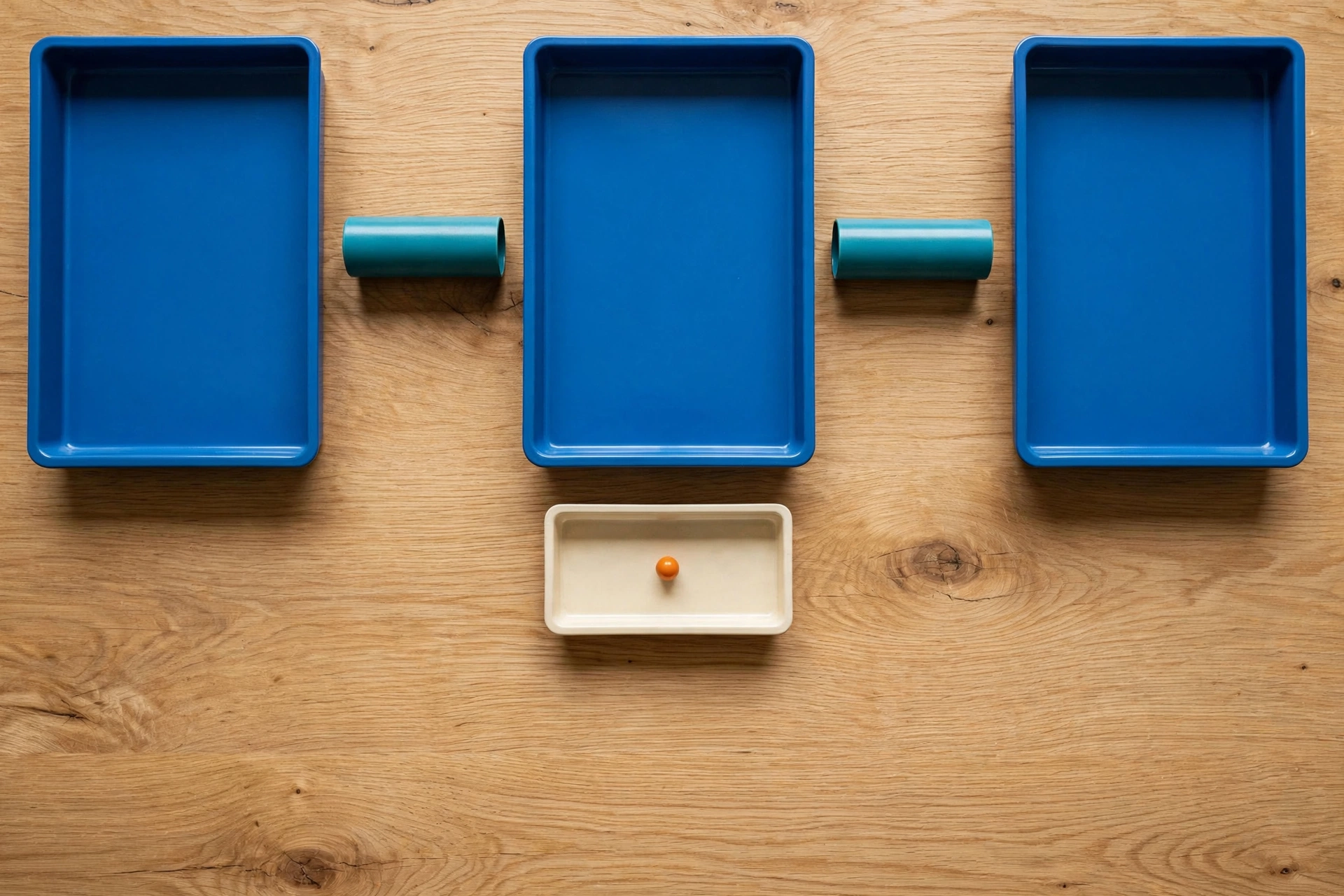 Three blue trays and two teal cylinders are arranged on a wooden surface, with an ivory tray containing an orange bead below.
