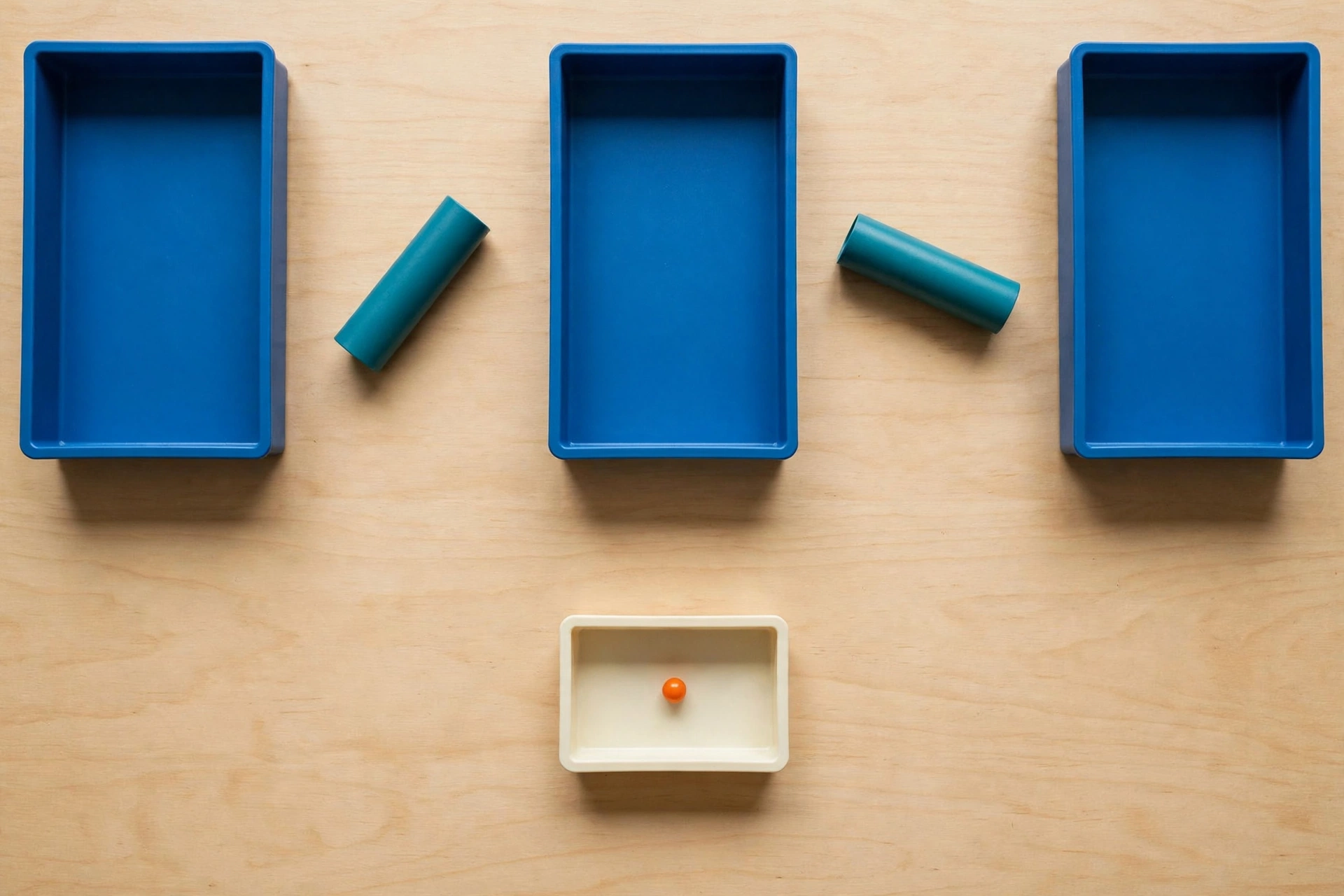 Three blue trays and two teal cylinders are arranged on a wooden surface, with an ivory tray containing an orange bead below.