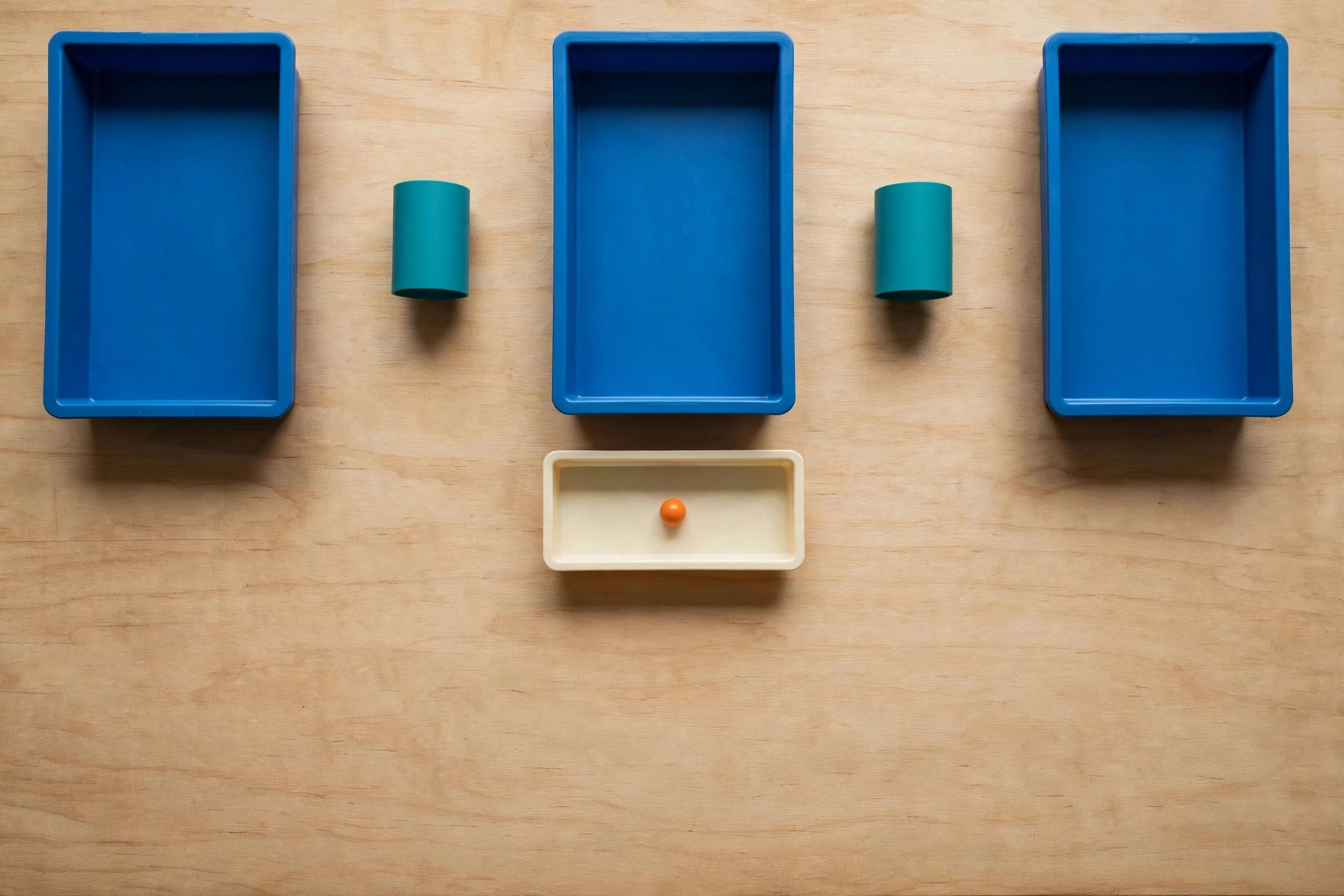Three blue trays and two teal cylinders are arranged on a wooden surface, with an orange bead in a lower ivory tray.