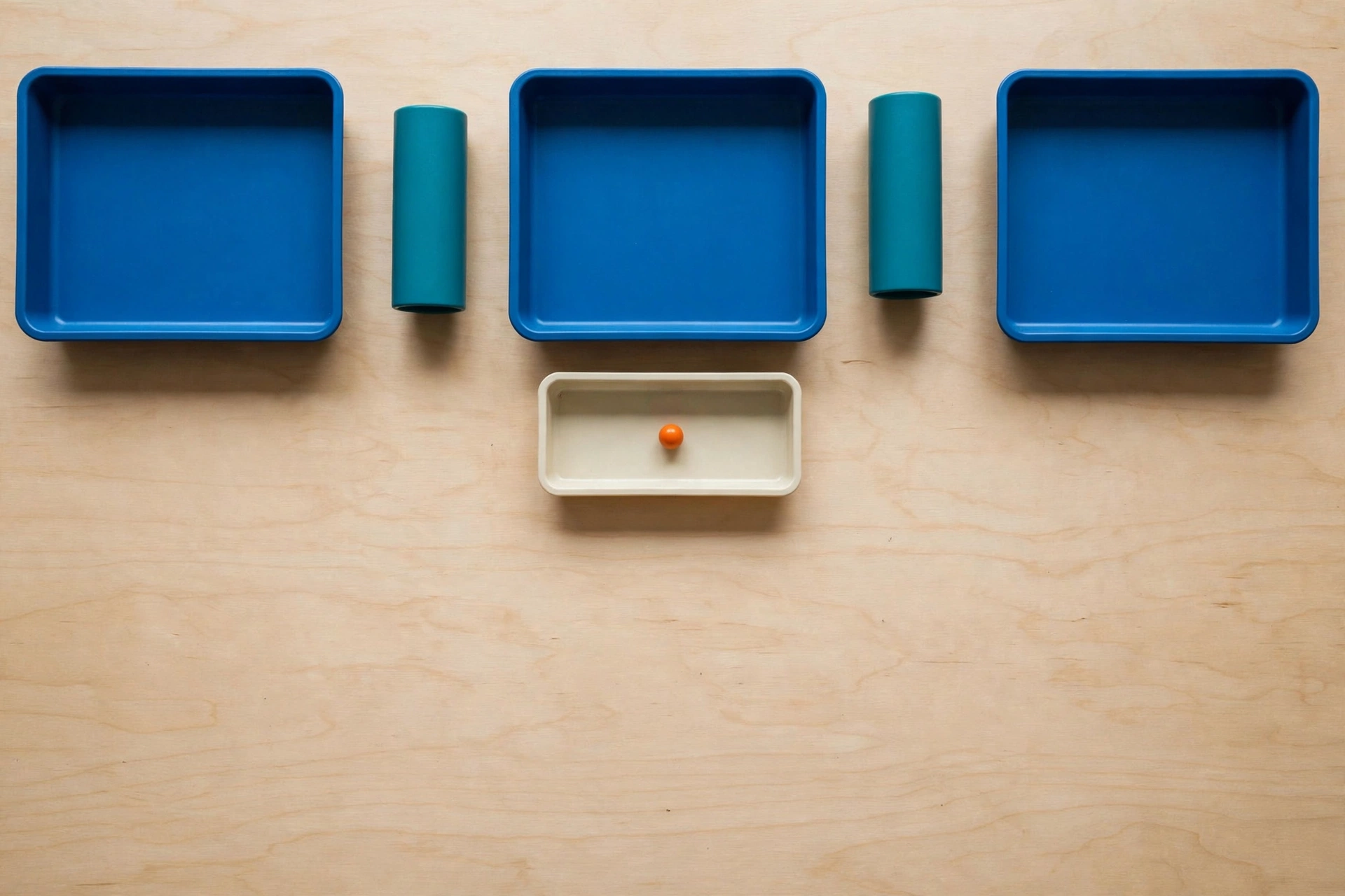 Three blue trays and two teal cylinders are arranged on a wooden surface, with a smaller ivory tray holding an orange bead below.