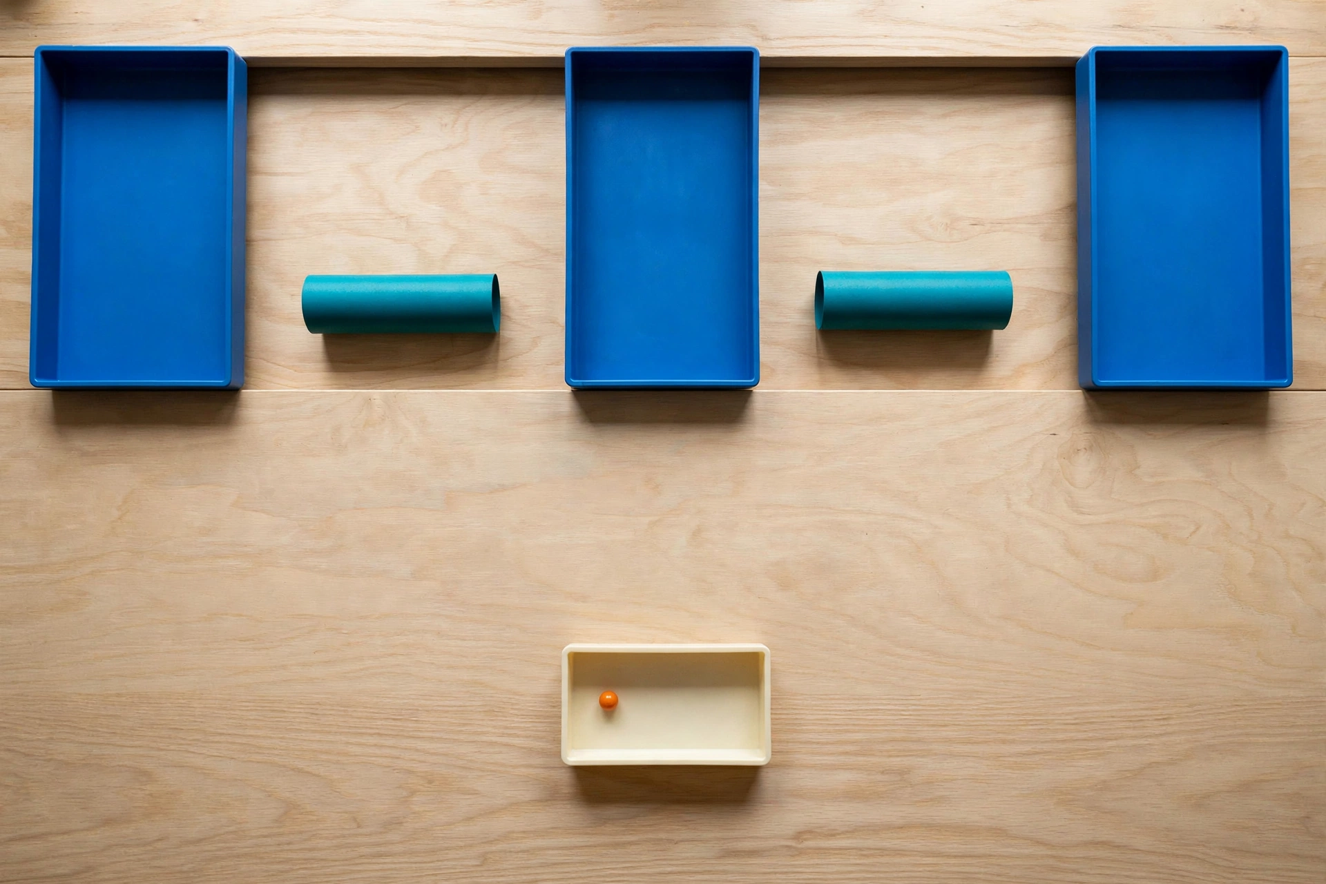 Three blue trays and two teal cylinders are arranged on a wooden surface, with an orange bead in a lower ivory tray.