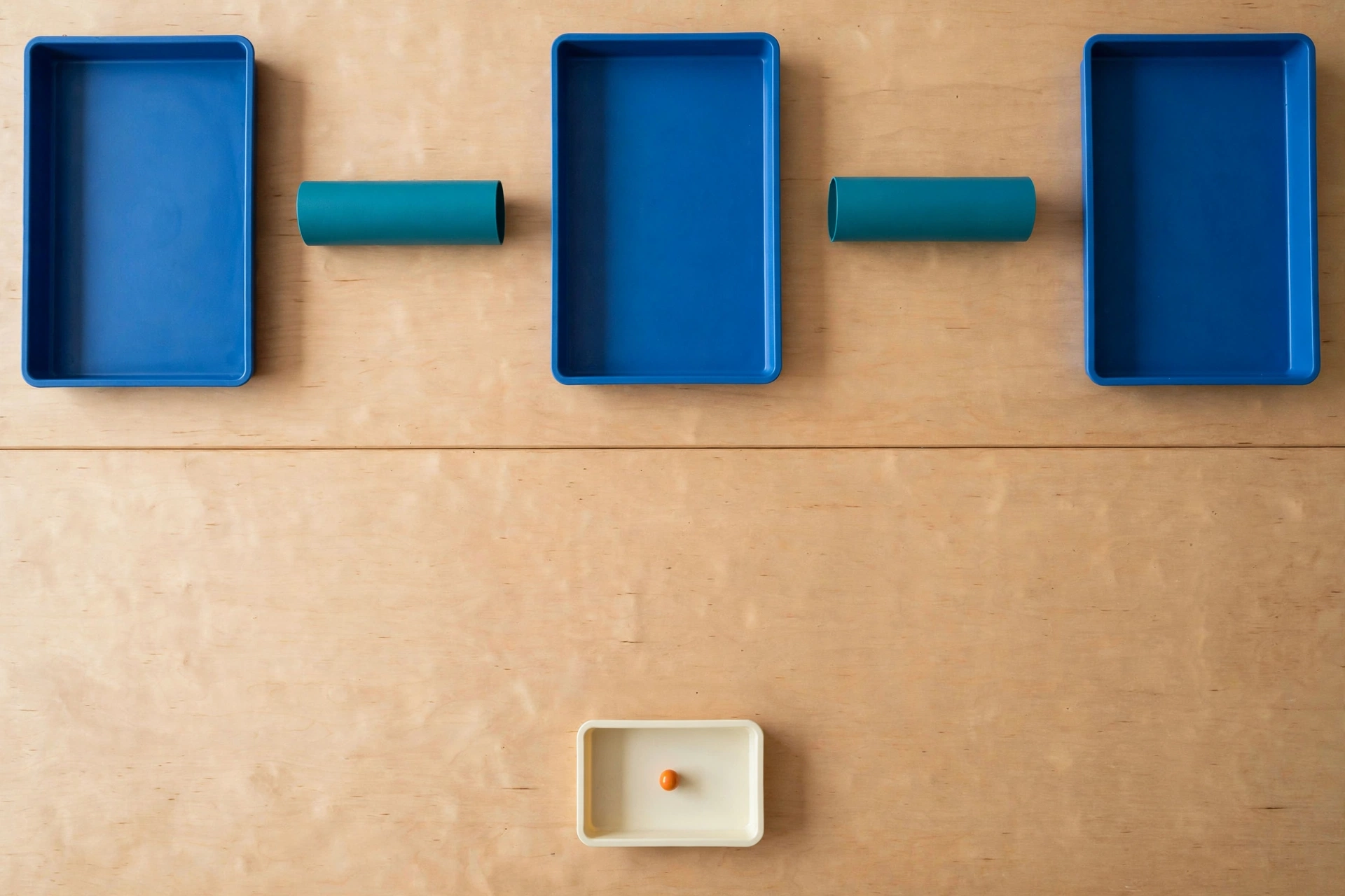 Three blue trays and two teal cylinders are arranged on a wooden surface, with a smaller ivory tray containing an orange bead below.