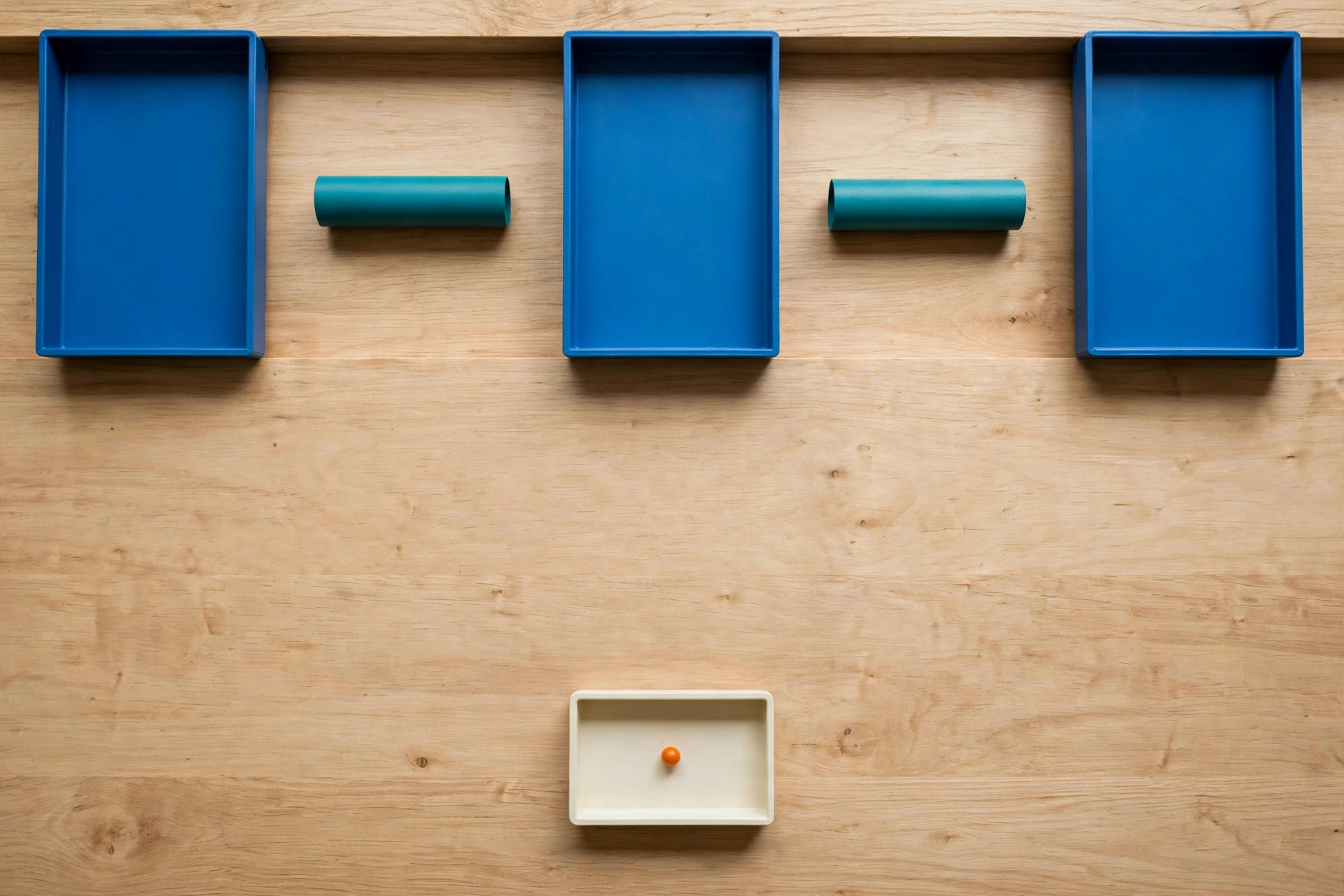 Three blue trays and two teal cylinders are arranged on a wooden surface, with a small ivory tray containing an orange bead below.