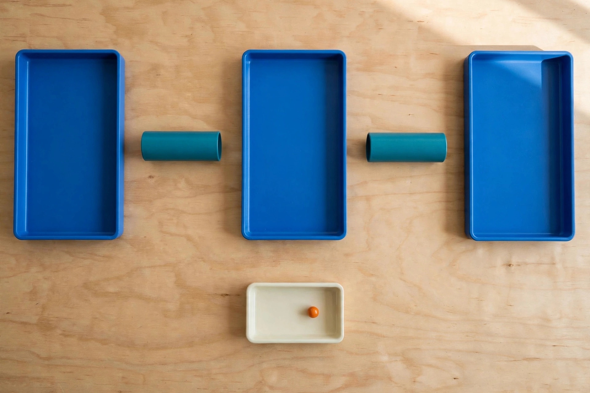 Three blue trays and two teal cylinders are arranged on a wooden surface, with an orange bead in a smaller ivory tray below.