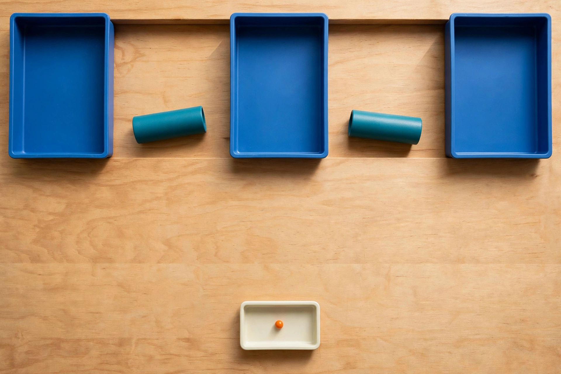 Three blue trays and two teal cylinders are arranged on a wooden surface, with a small ivory tray holding an orange bead below.