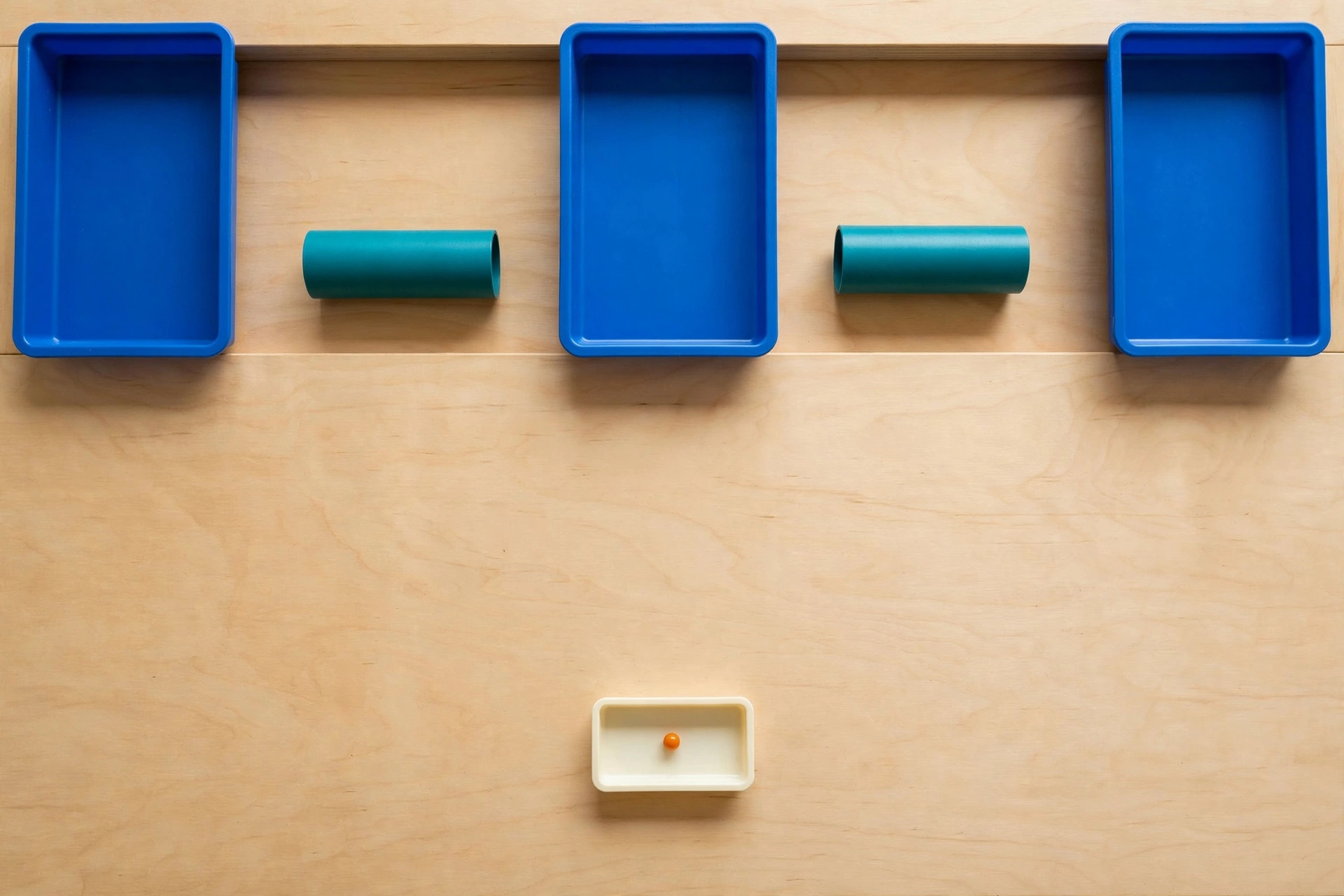 Three blue trays and two teal cylinders are arranged on a wooden surface, with a small ivory tray containing an orange bead below.