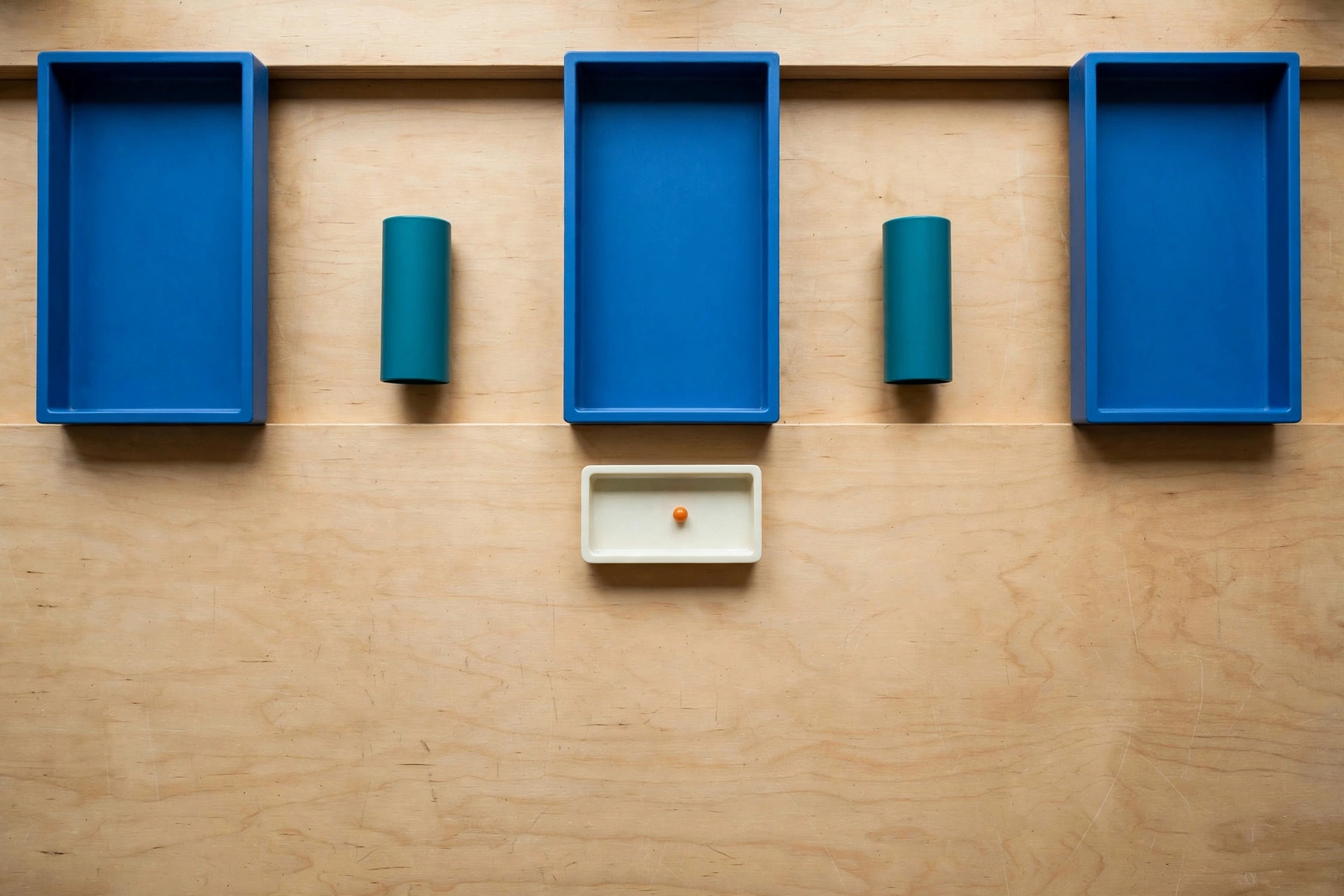 Three blue trays and two teal cylinders are arranged on a wooden surface, with a small ivory tray containing an orange bead below.