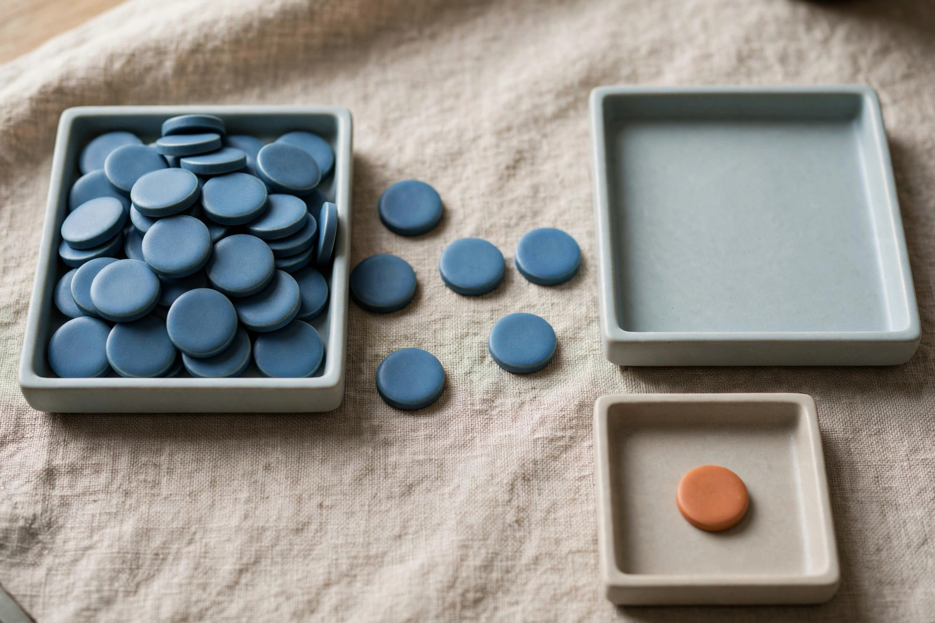 Three shallow trays hold blue ceramic tokens, with one tray containing an orange token, on a textured cloth surface.
