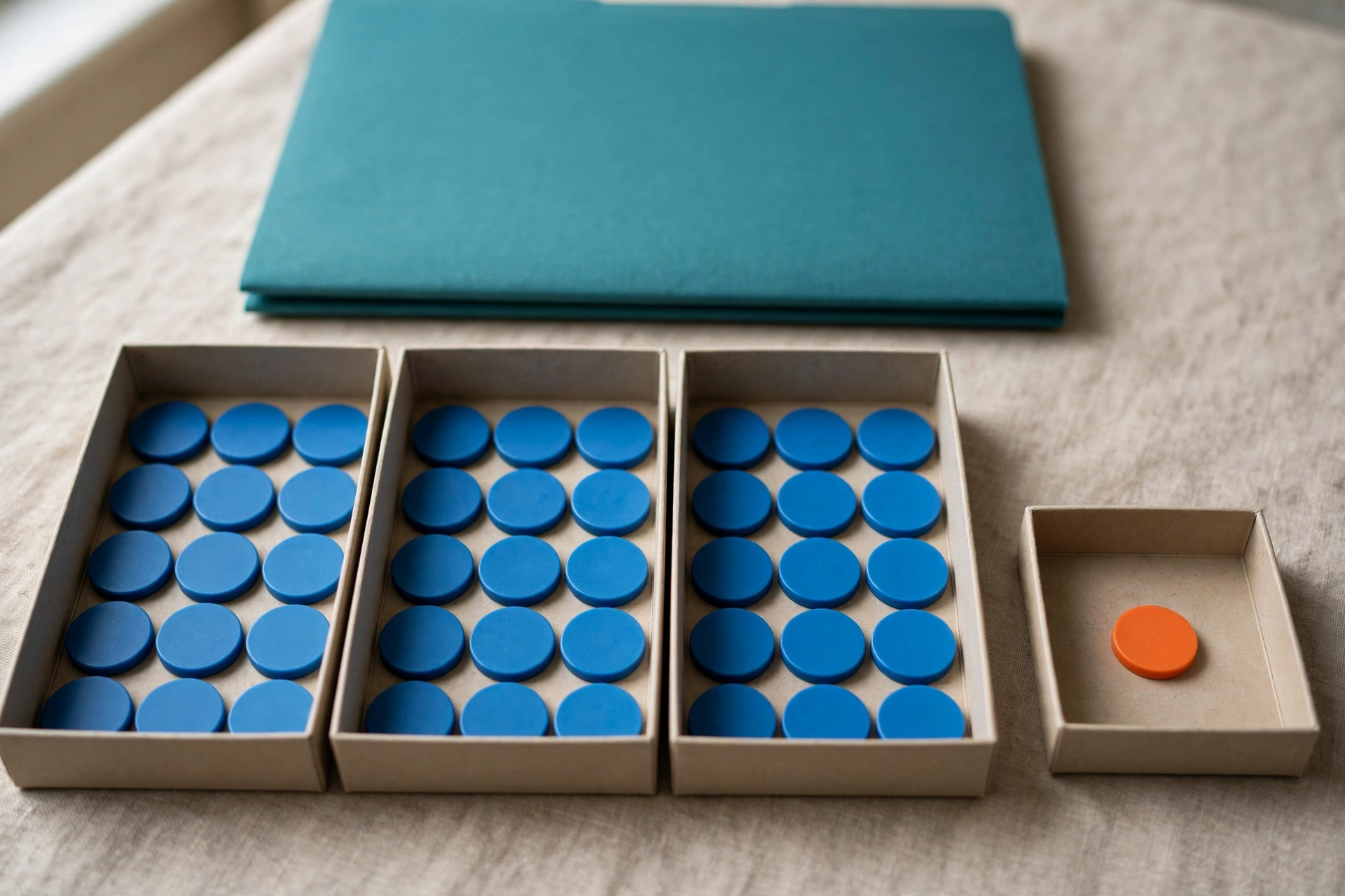 Three trays hold blue tokens, and a fourth tray holds a single orange token, with a teal folder behind them on a textured surface.