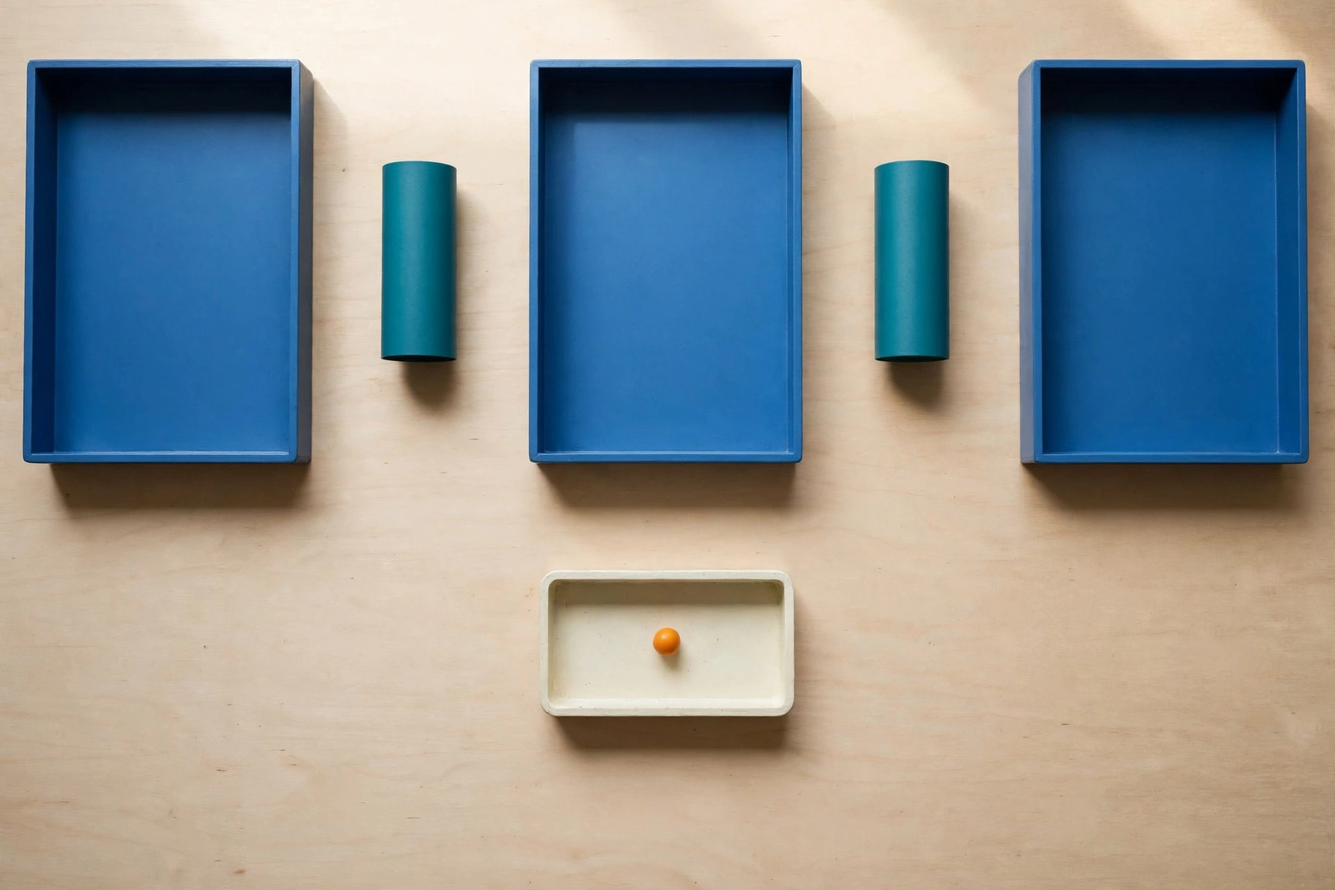 Three blue trays and two teal cylinders are arranged on a light wooden surface, with an ivory tray containing an orange bead below.