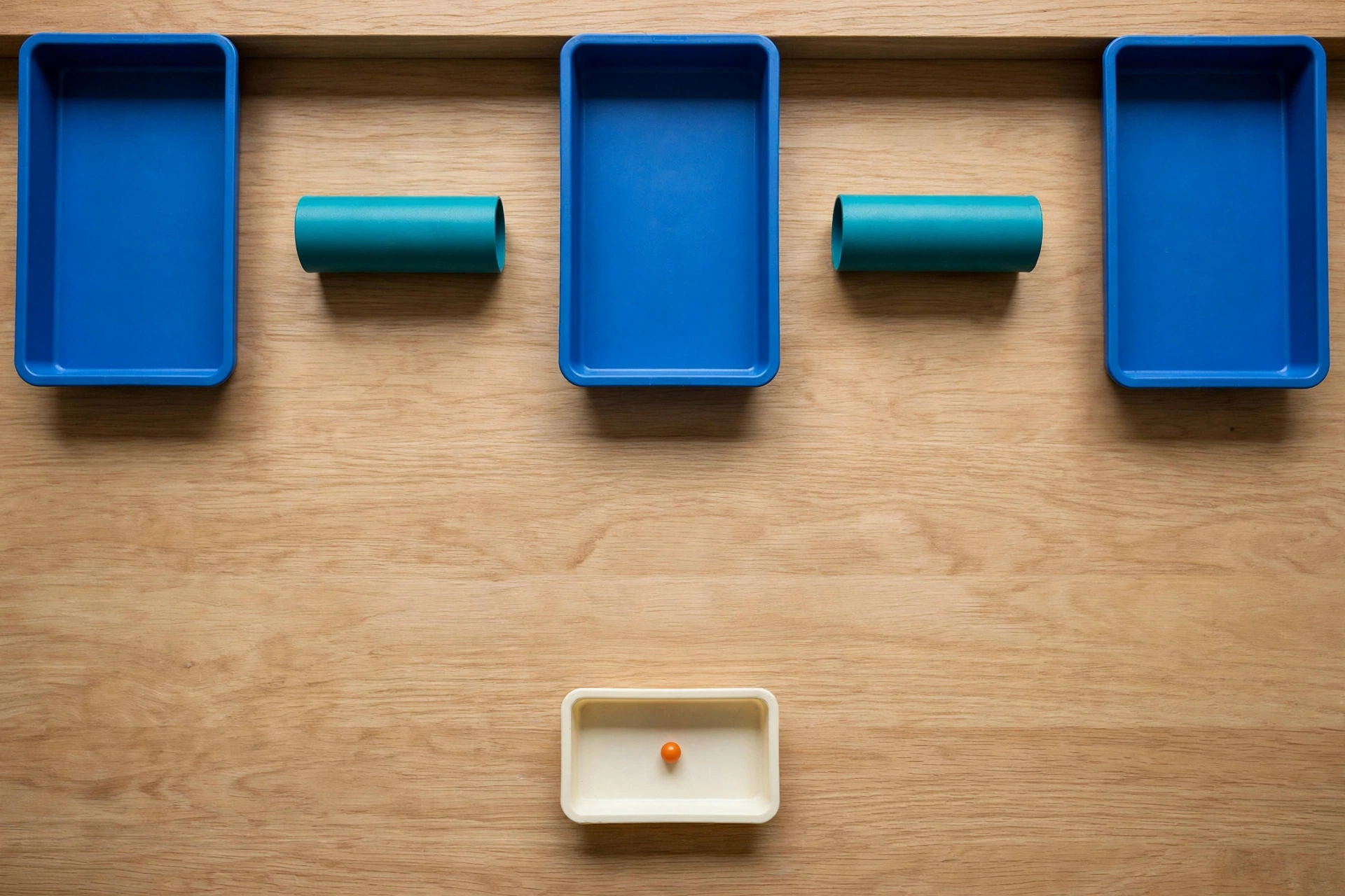 Three blue trays and two teal cylinders are arranged on a wooden surface, with a small orange bead in a separate ivory tray below.