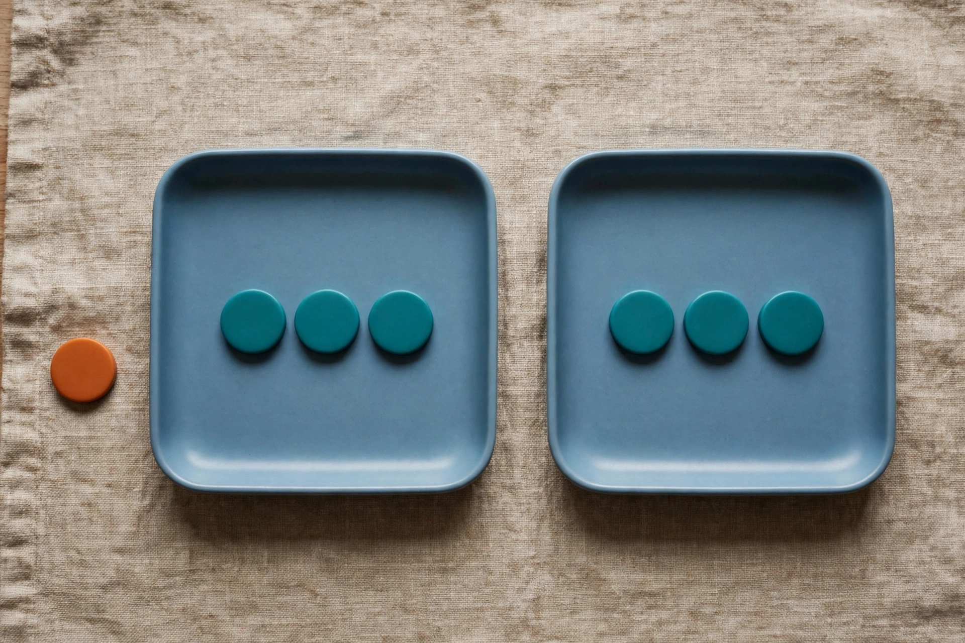 Two blue trays each hold three teal tokens, with one orange token beside the left tray on a textured surface.