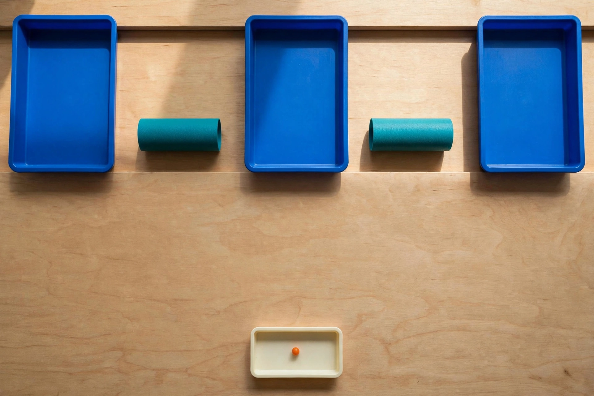 Three blue trays and two teal cylinders are arranged on a wooden surface, with a small orange bead in a lower ivory tray.