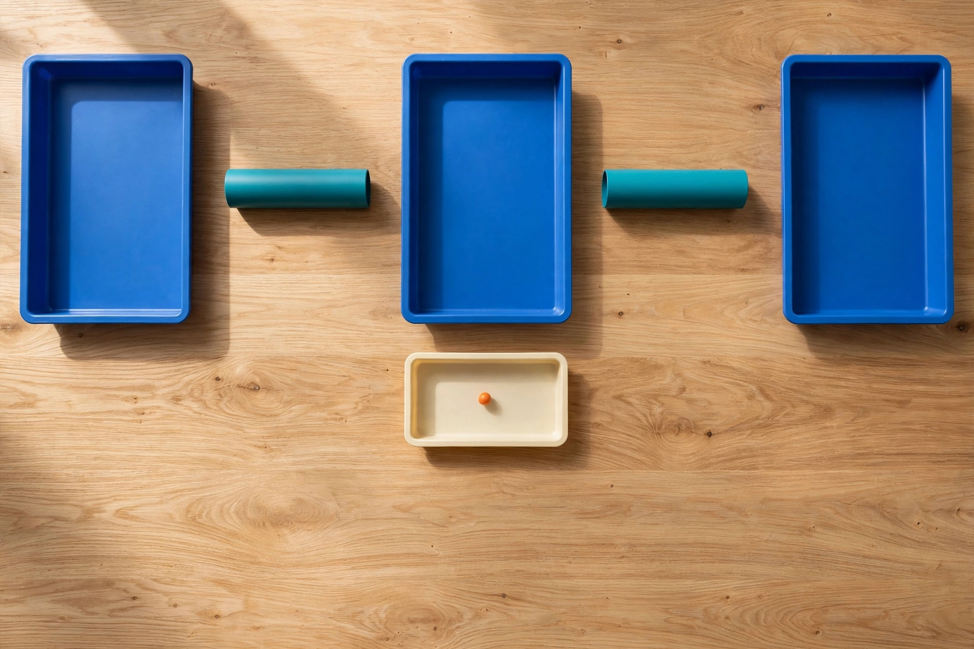 Three blue trays and two teal cylinders are arranged on a wooden surface, with a smaller ivory tray containing an orange bead below.