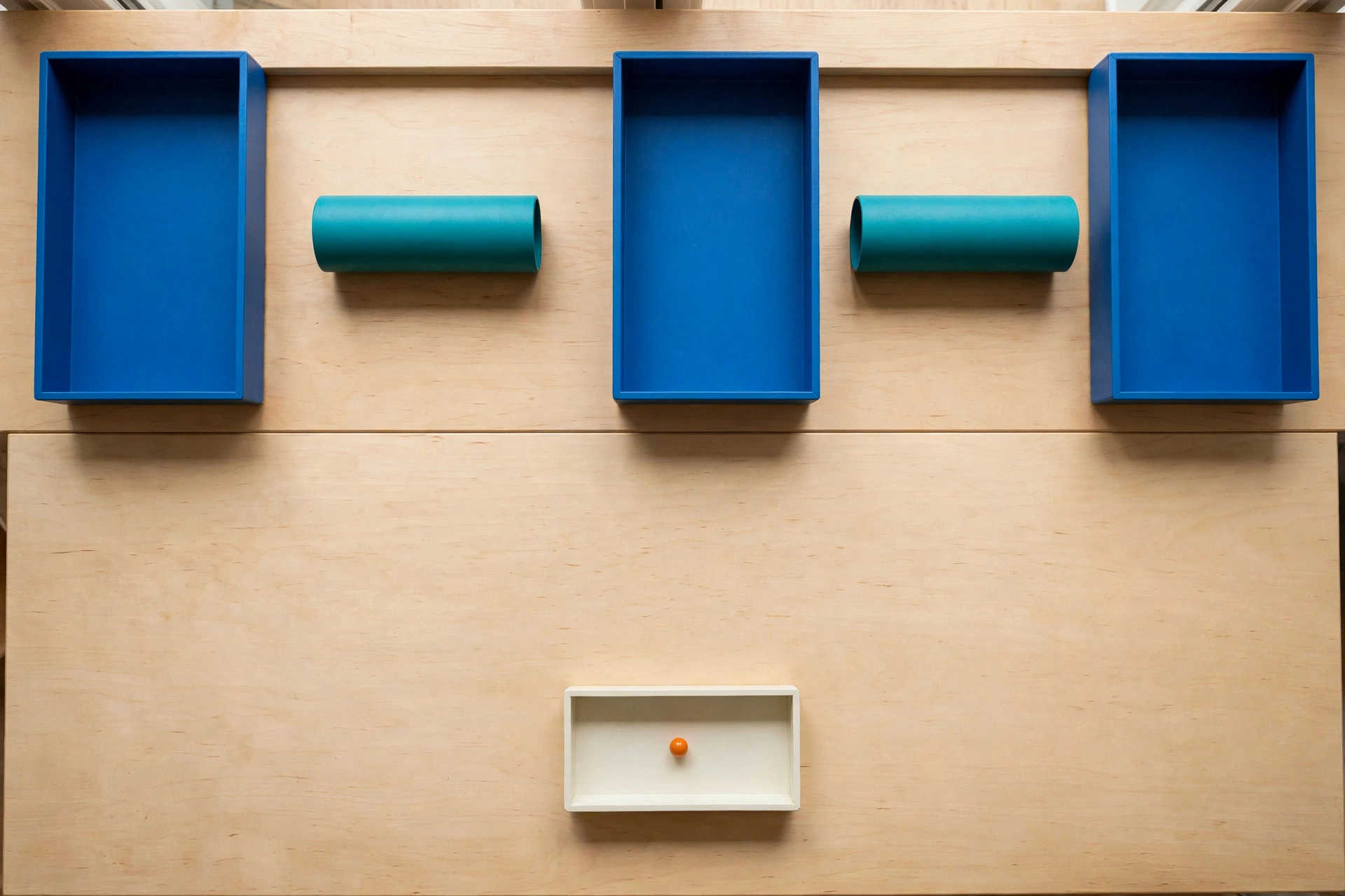 Three blue rectangular trays are arranged on a light wood surface with two teal cylinders in the gaps between them.