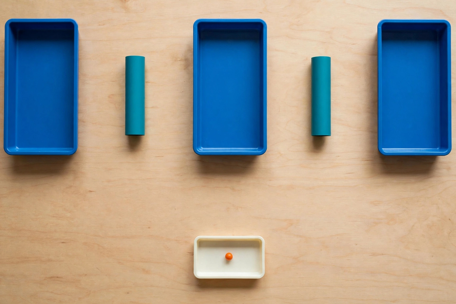 Three blue trays and two teal cylinders are arranged on a wooden surface, with an orange bead in a smaller ivory tray below.